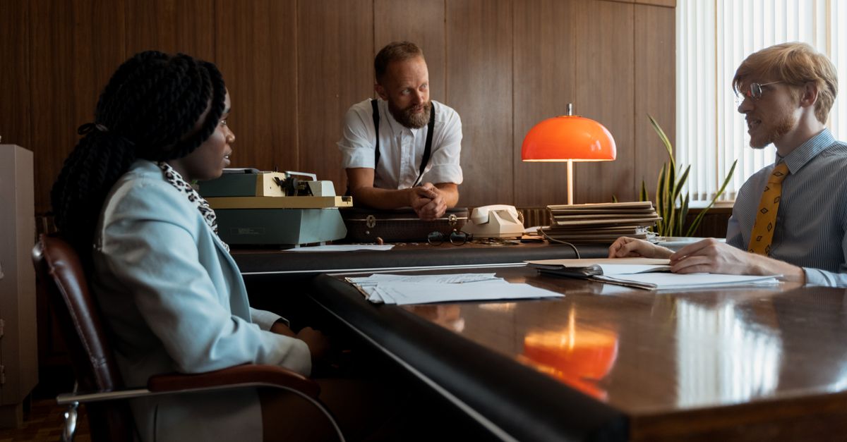Three colleagues engage in a discussion around a vintage desk in a retro office space.