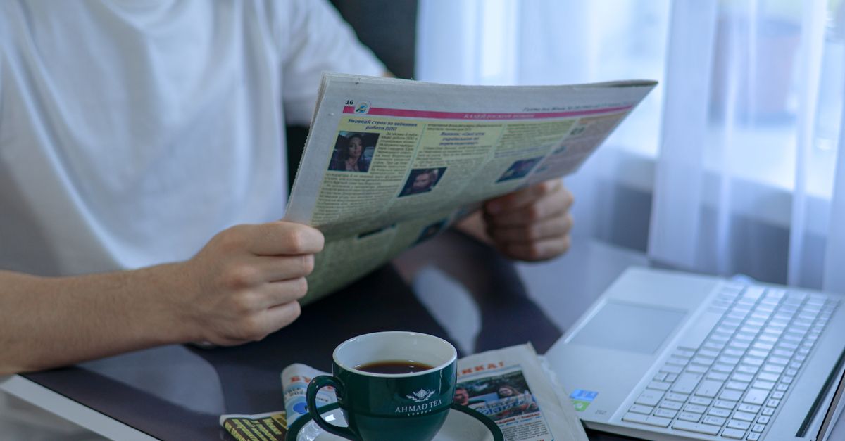 Adult enjoying coffee and newspaper at a desk with laptop. Indoors, bright daylight setting.