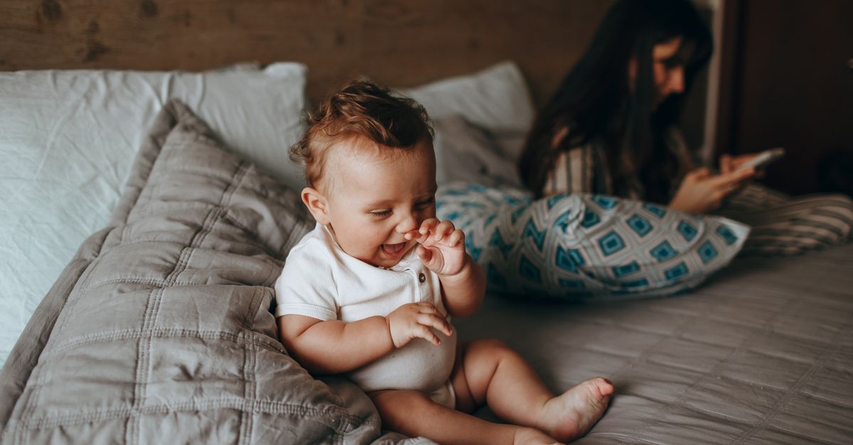 Adorable baby plays happily on a cozy bed while mother relaxes nearby, creating a warm family moment.