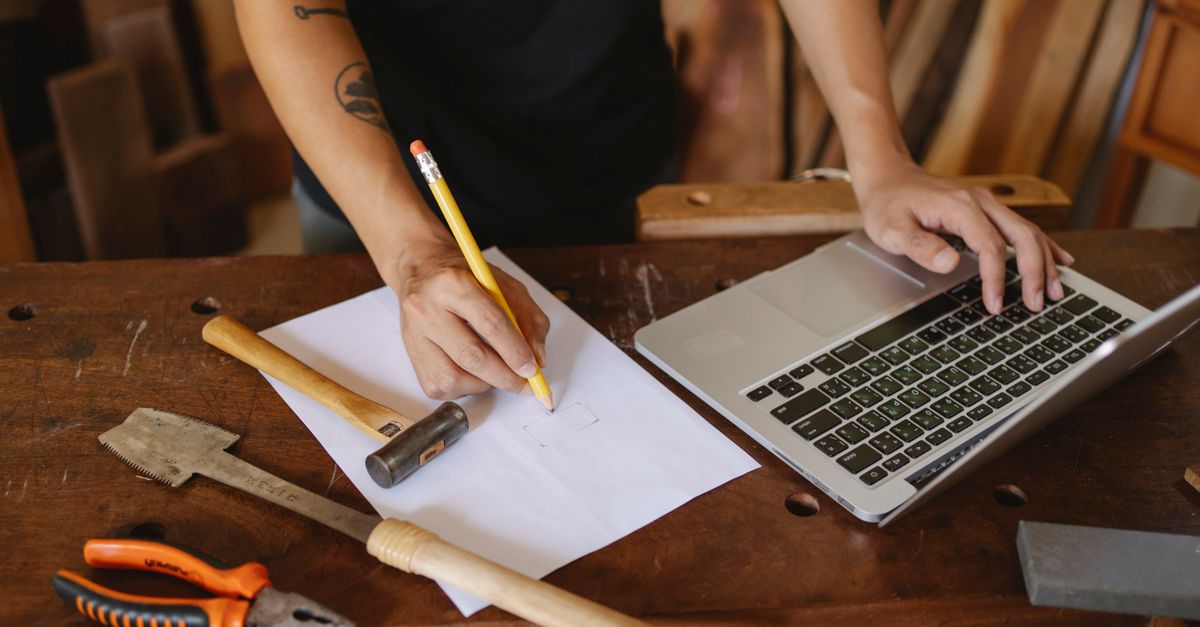 Crop faceless male worker standing near table and creating new craft Piece on paper while working with laptop
