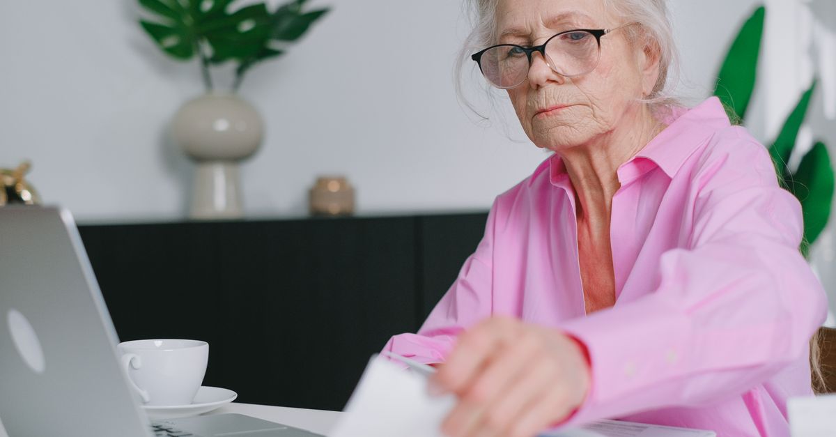 Elderly woman in pink shirt working on a laptop at home, focused and engaging with documents.