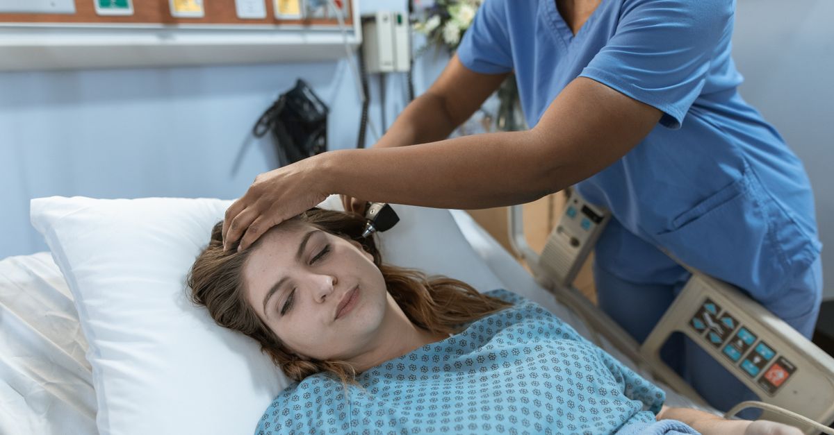 Crop African American female doctor with professional equipment doing examination of ear of woman lying on bed in hospital ward