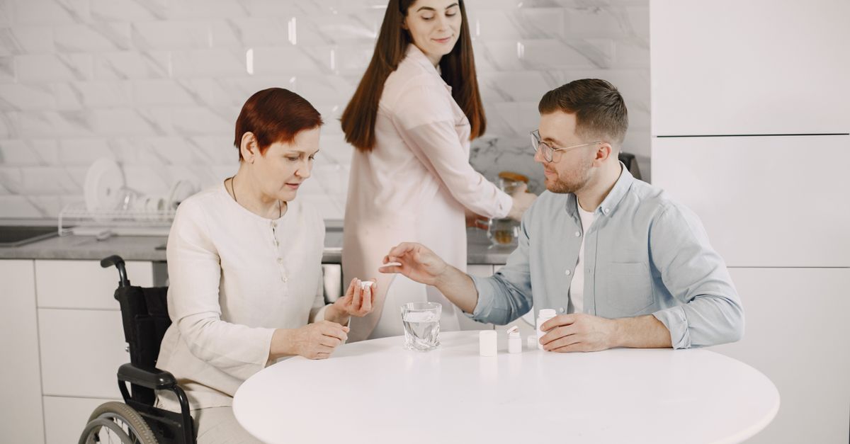 Elderly woman in wheelchair receiving help with medication from caregiver in modern kitchen.