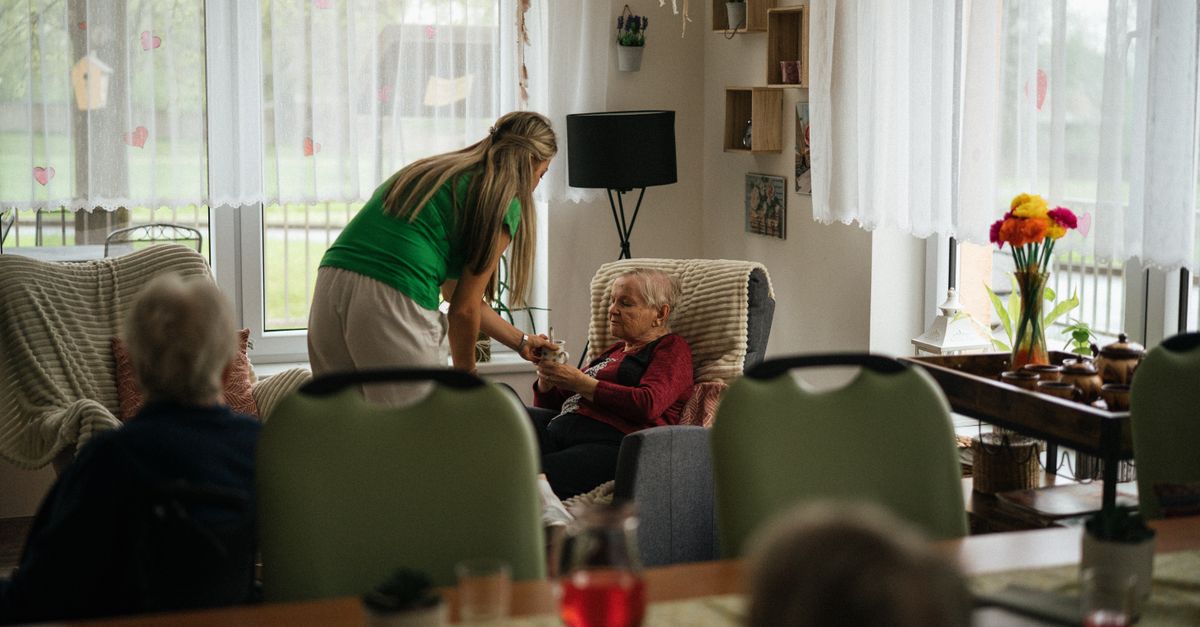 Caregiver assisting elderly women with tea in a cozy nursing home setting.